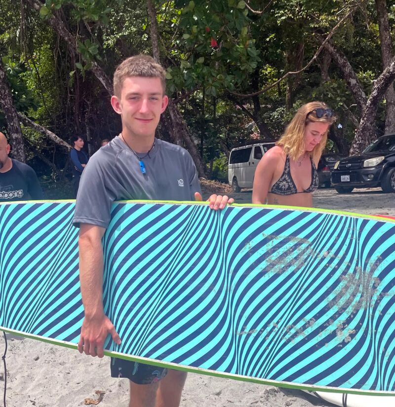 A young man stands on a beach, holding a blue and white striped surfboard. He is wearing a gray t-shirt and dark shorts. Behind him, a woman in a bikini top stands near a parked car. The background includes trees and other people, suggesting a beach setting. The man is looking at the camera with a slight smile.